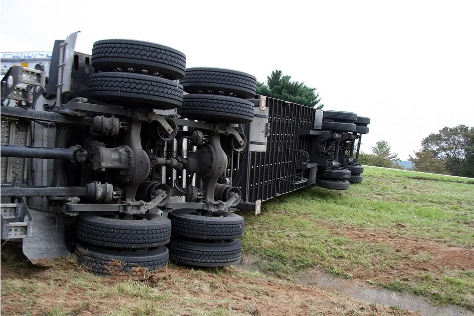 A large truck or semi flipped over in the grass near a highway